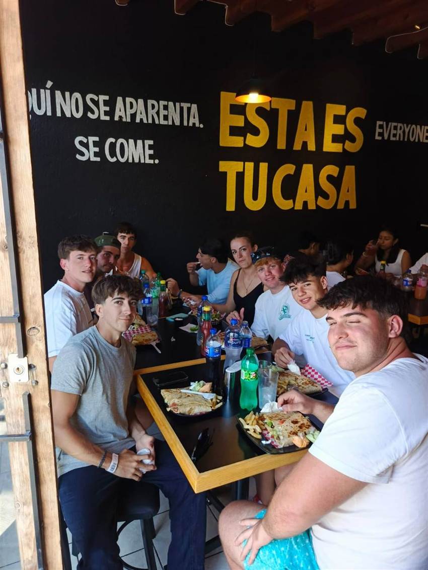 Un grupo de jóvenes viajeros comiendo tortas y burritos bajo el mural "Esta es tu casa"